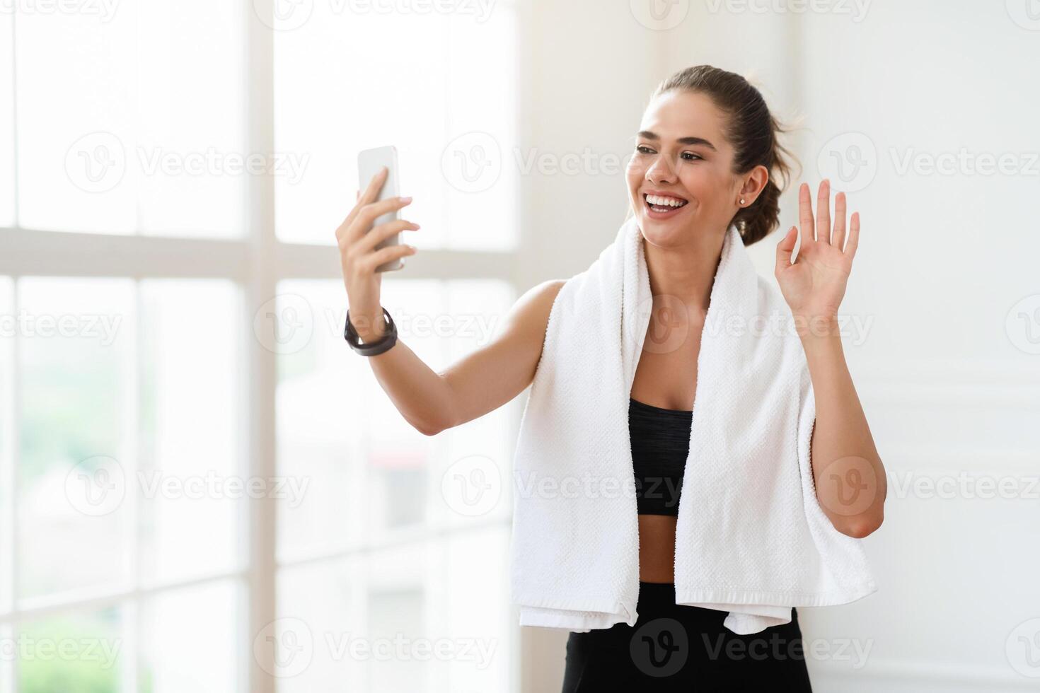Athletic young woman with towel having online conference with fitness trainer or recording blog. Sporty pretty lady waving at smartphone screen, greeting her instructor or followers, free space photo