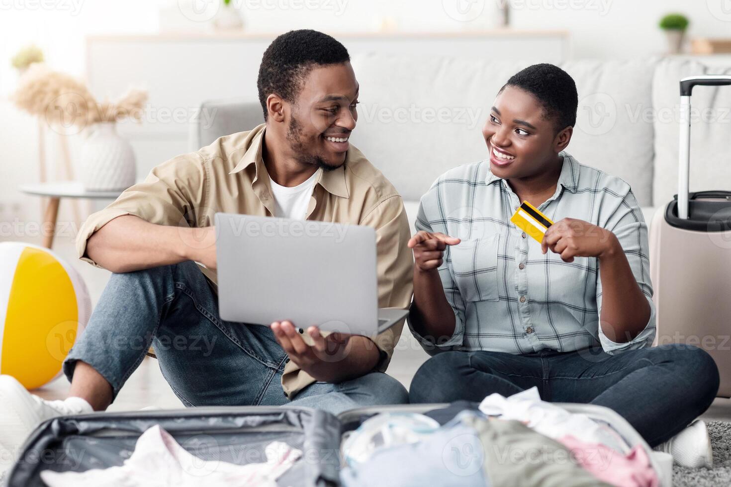 Cheerful Black Tourists Couple Booking Tickets Online With Laptop And Credit Card Sitting Near Unpacked Suitcase At Home. Book Your Tour, Traveling Website And Internet Services. Selective Focus photo