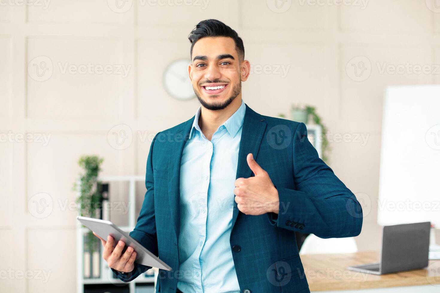 Smiling Arab businessman holding computer, showing thumb up gesture in office. Handsome guy entrepreneur recommending online services, having virtual conference with colleagues photo