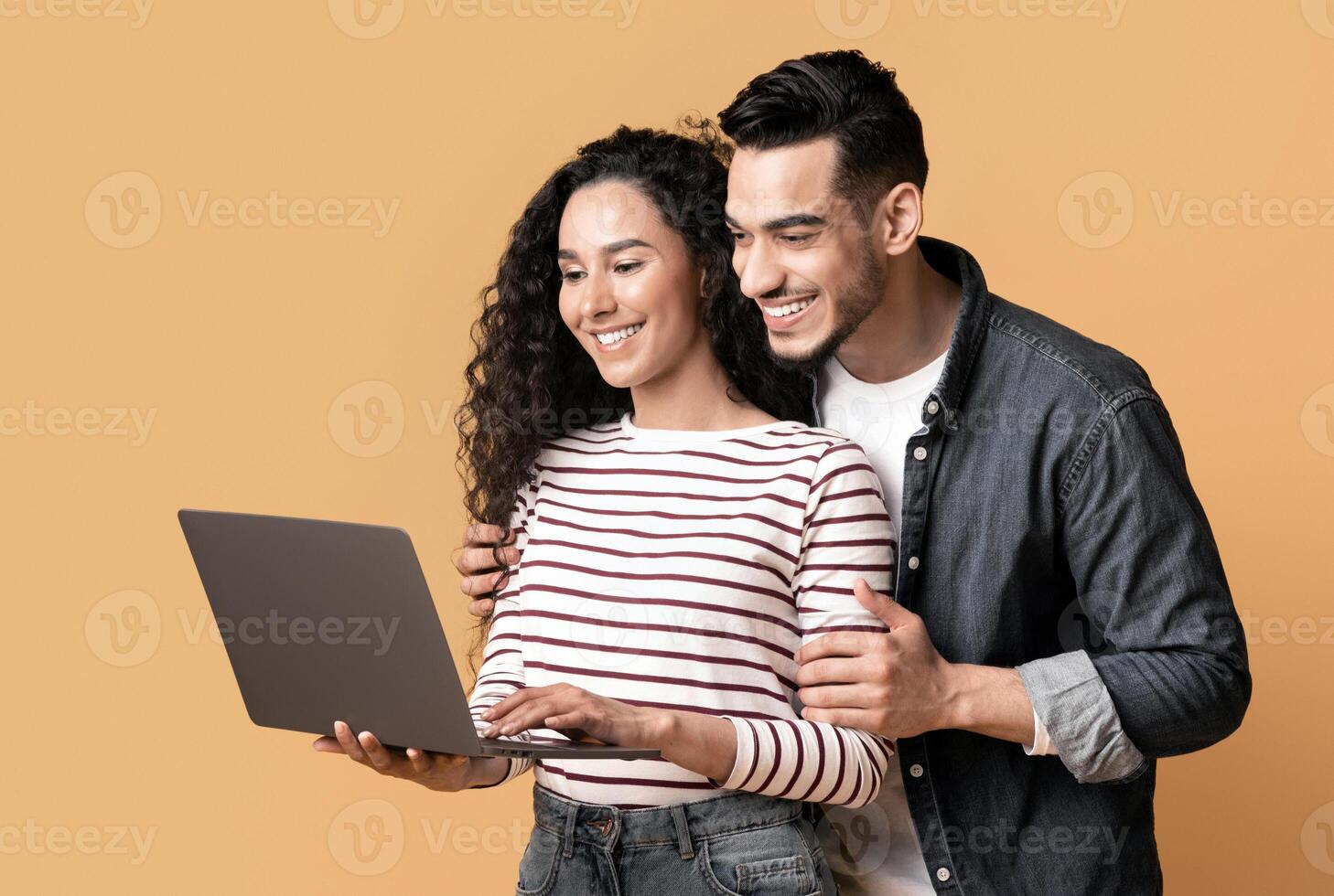 Young middle eastern couple using laptop together while standing over yellow background in studio, cheerful arab spouses shopping online or browsing new website on computer, panorama with copy space photo