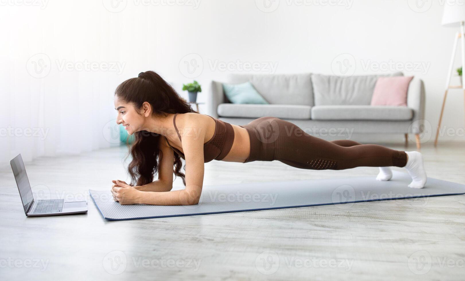 Sporty Indian woman standing in elbow plank, following tutorial on laptop at home. Side view of Asian lady exercising her abs core muscles. Online trainings during covid lockdown photo