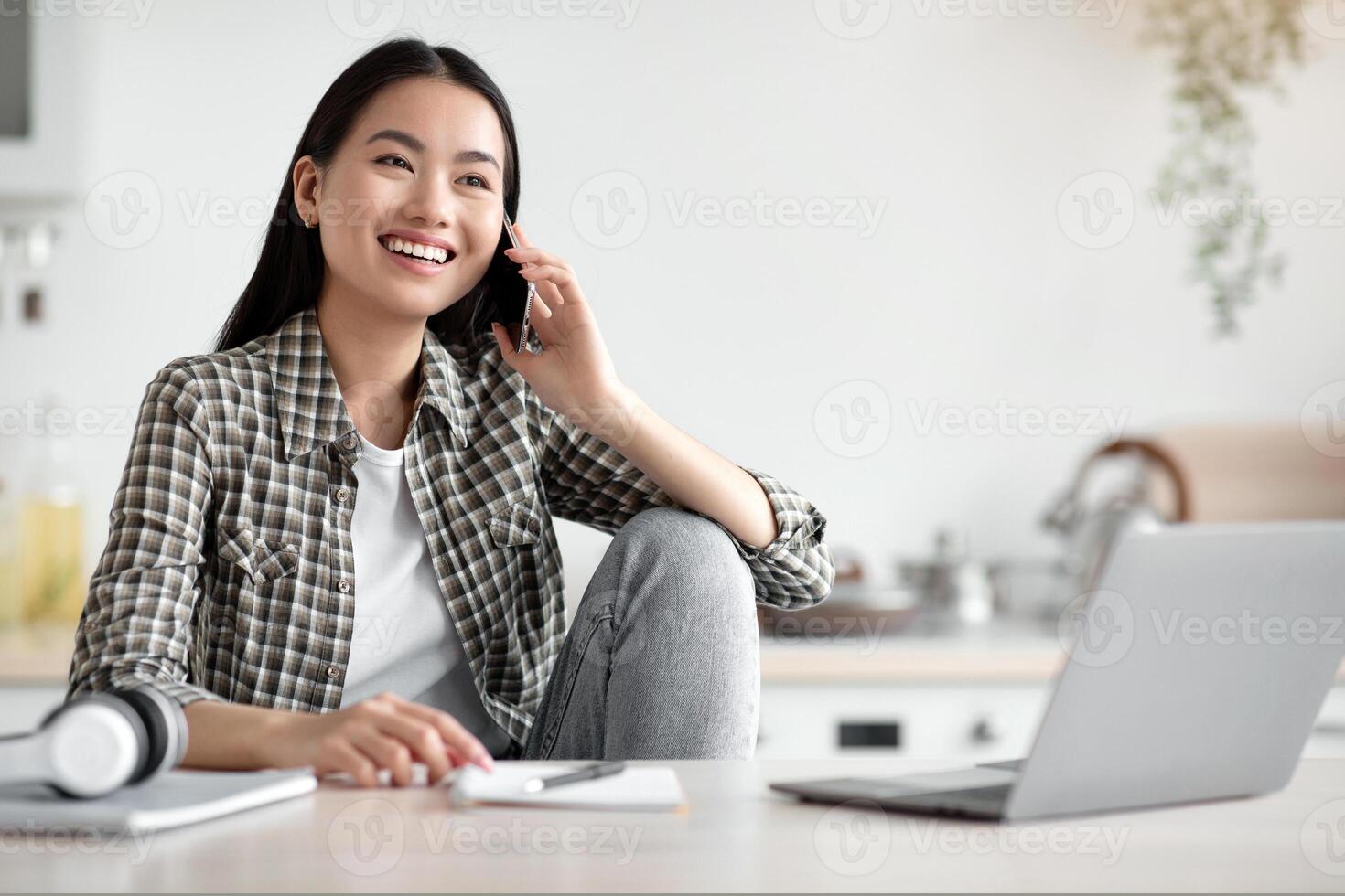 Smiling young asian woman freelancer talking on mobile phone and using laptop, working from home during isolation, looking at copy space. Happy lady having conversation on phone at kitchen photo