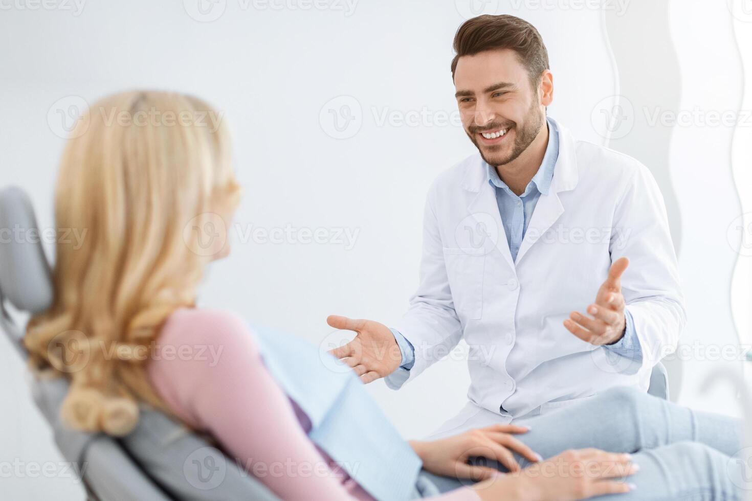 Friendly dentist handsome millennial man in workwear having conversation with blonde woman patient sitting at dental chair, smiling and gesturing, side view. Doctor and patient relationship photo