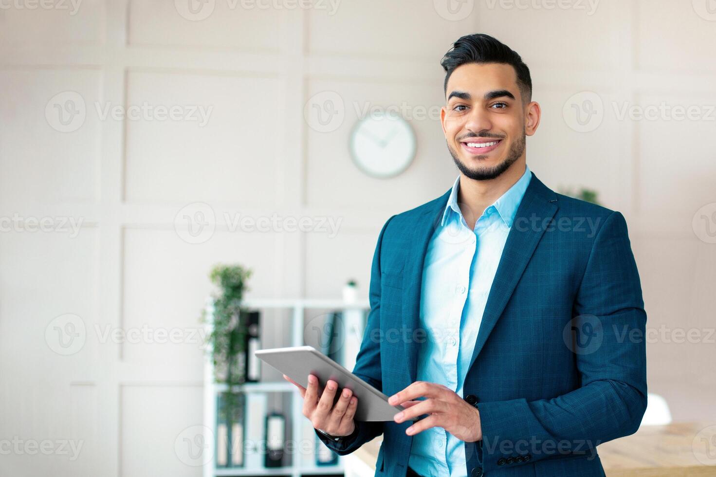 Happy guy office worker in formal suit holding tablet computer and smiling at camera in modern office. Young Arab manager checking email, searching for business info on web photo