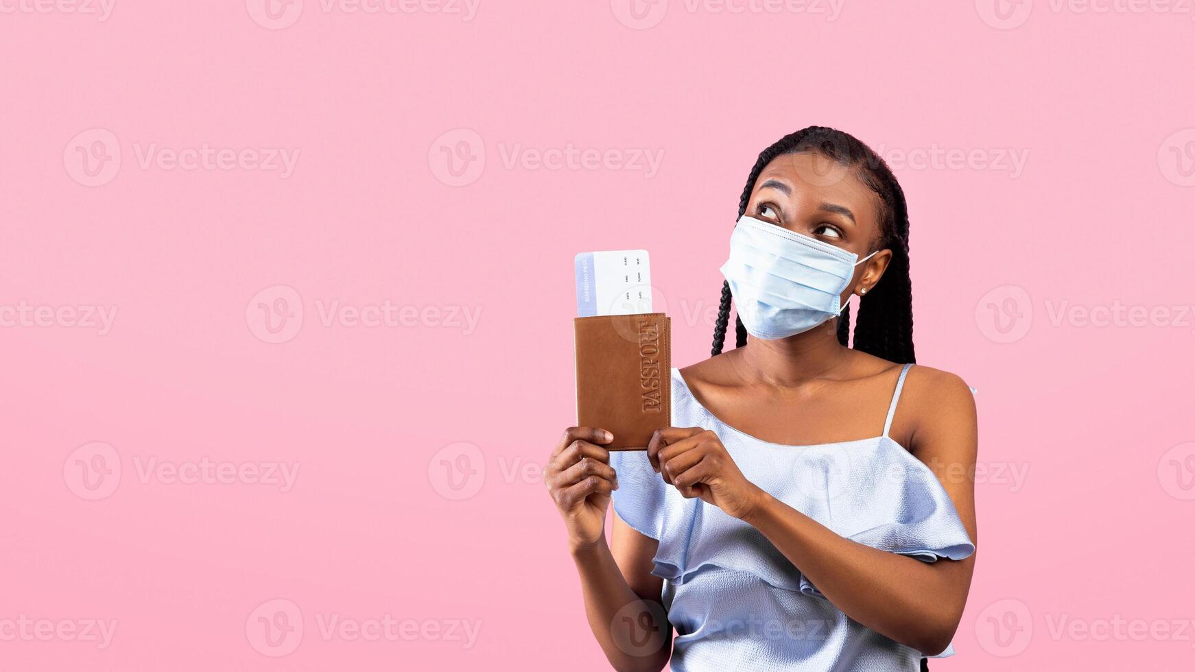 Millennial black woman wearing face mask, holding international passport and plane tickets, looking aside on pink studio background. Traveling and tourism during coronavirus epidemic photo