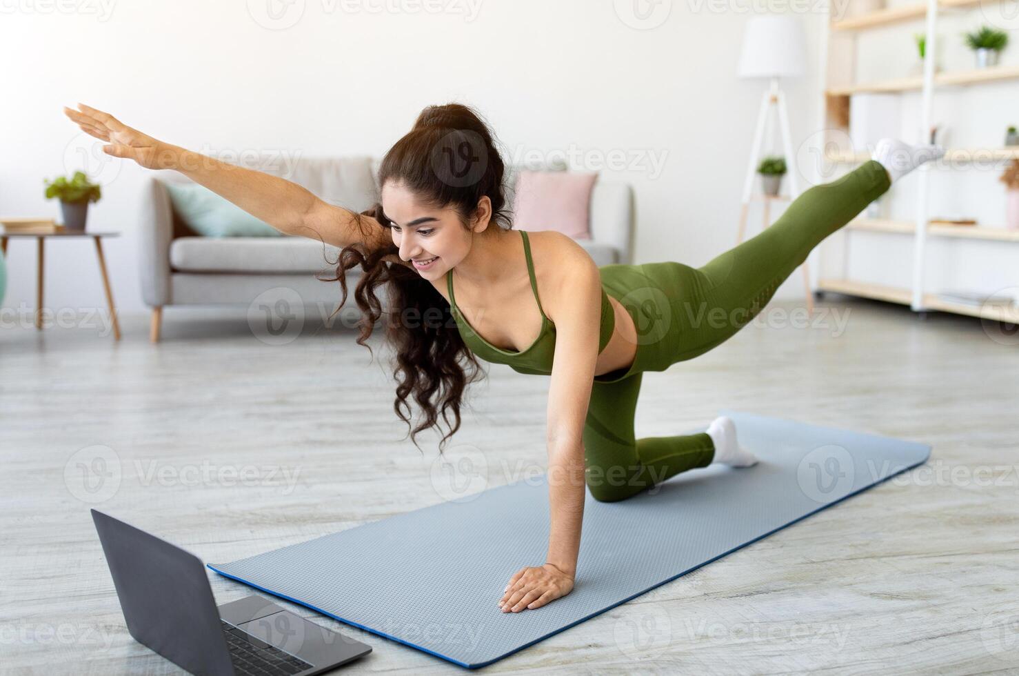 Online sports during covid lockdown. Millennial Indian woman exercising on mat, watching sports on laptop at home. Active Asian lady leading active lifestyle, training her booty muscles photo