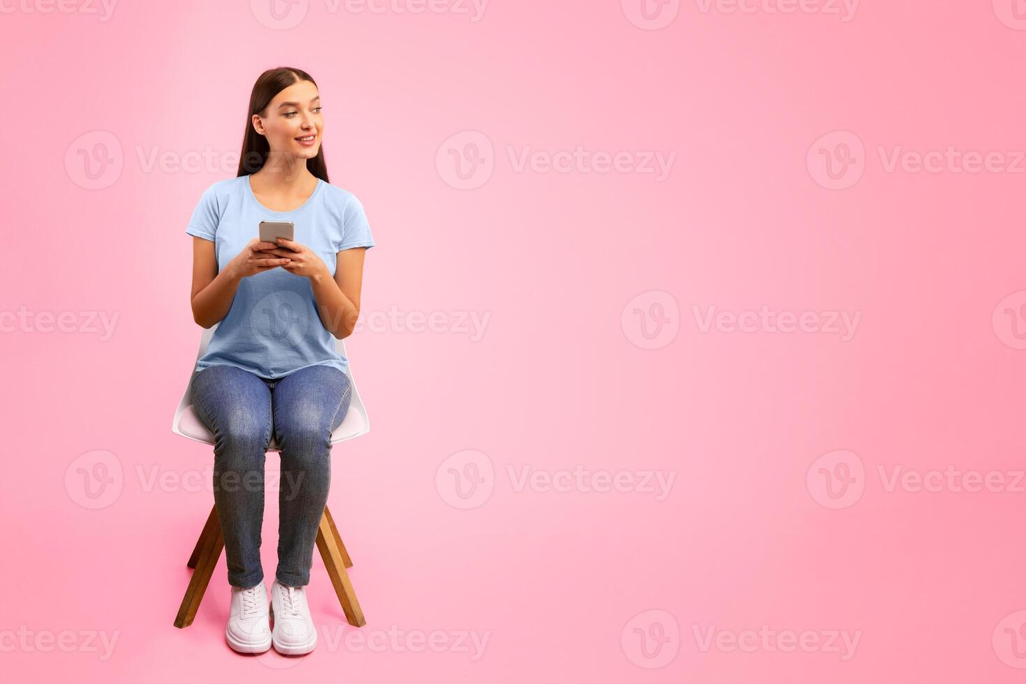 Portrait of curious girl sitting on the chair, using her cell phone and looking aside at copy space, orange wall photo