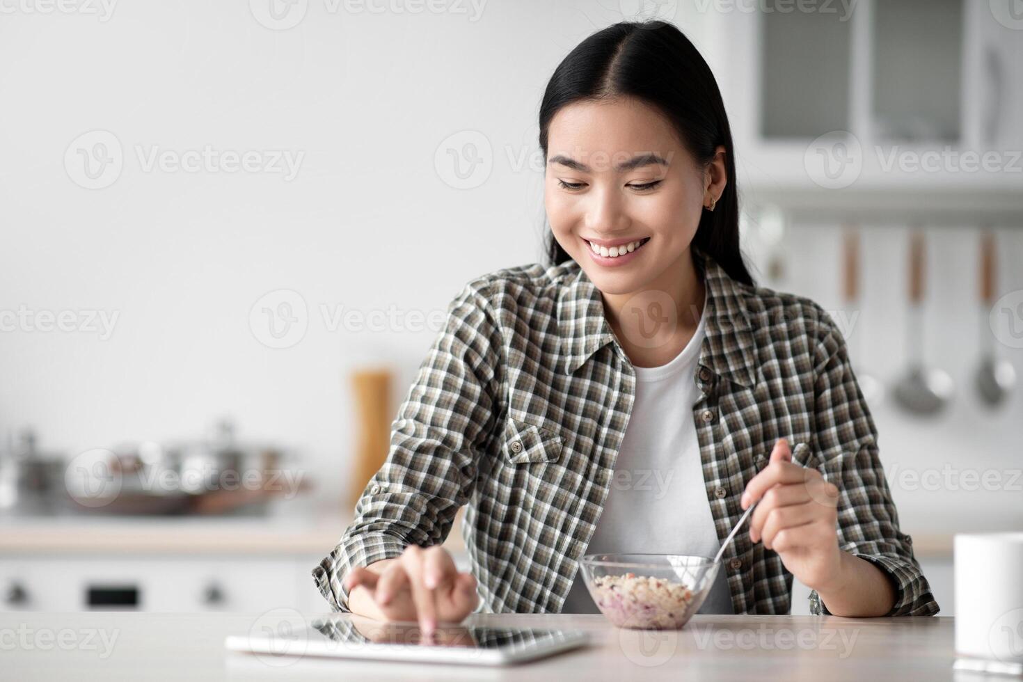 Smiling asian woman having breakfast and surfing on social media, using digital tablet, kitchen interior, copy space. Happy young lady eating oatmeal and reading blog or book on pad photo