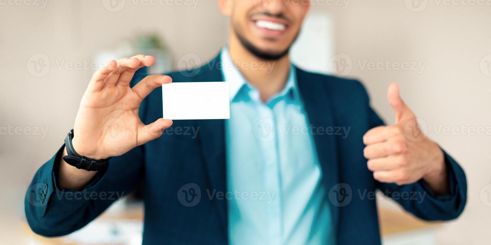 Unrecognizable Arab entrepreneur holding empty business card, gesturing thumb up at office, mockup for design. Cropped view of Eastern businessman demonstrating blank advertising card photo