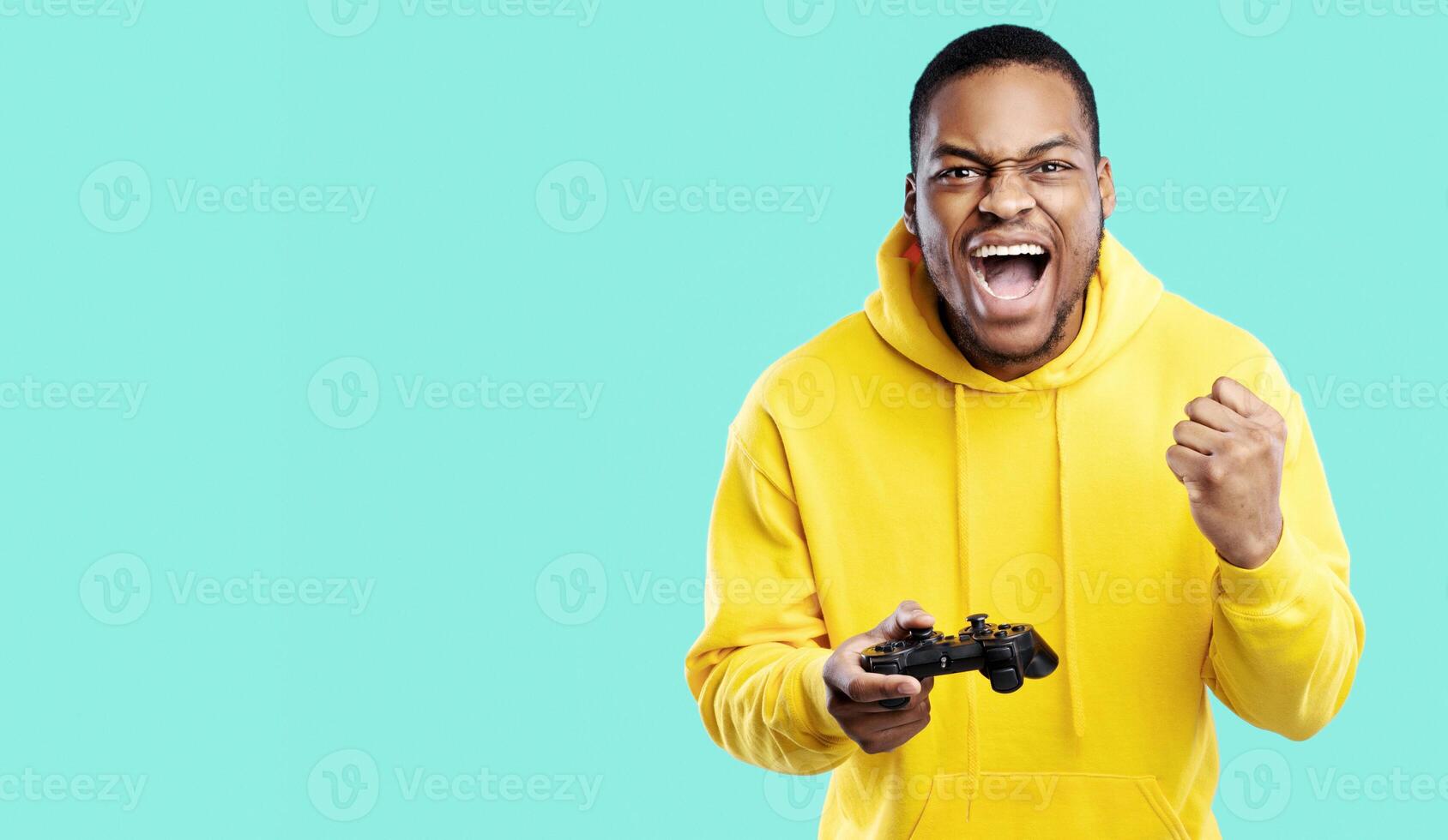 Gaming Concept. Emotional Black Guy Winning Game Posing With Joystick And Gesturing Yes In Joy Over Blue Studio Background. Millennial Man Celebrating Victory Playing Videogame. Panorama photo