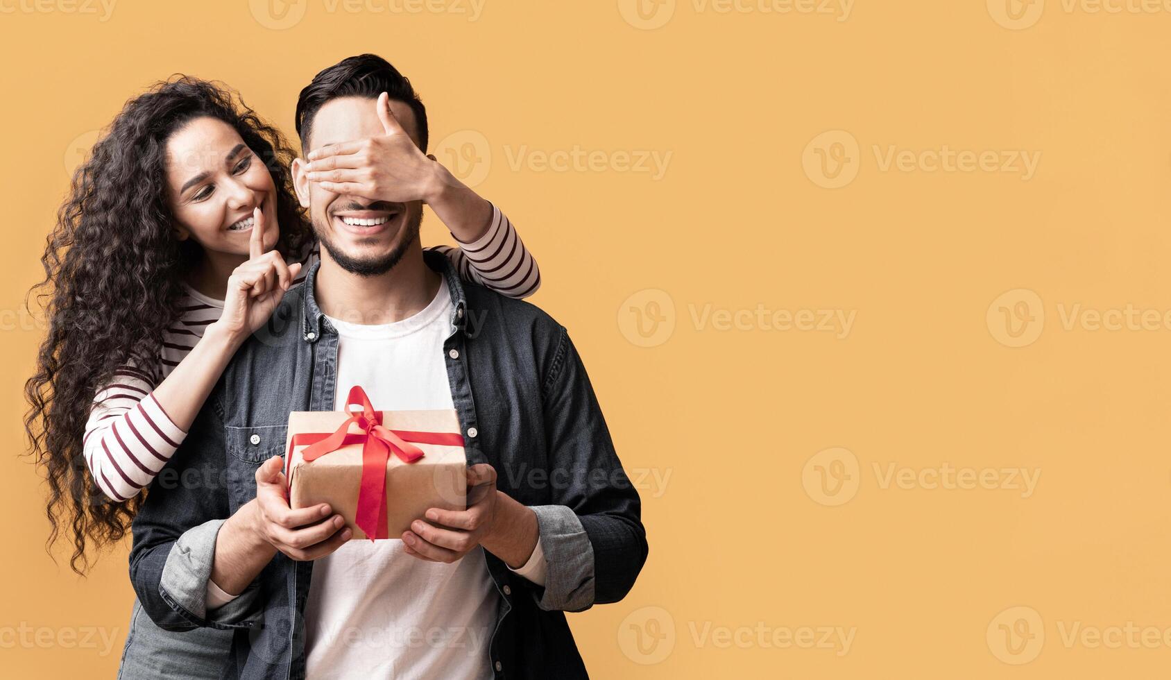 Surprise Present. Romantic young arab woman covering her boyfriend's eyes and giving gift, happy lady showing shh silence sign while surprising her lover, standing together on yellow background photo