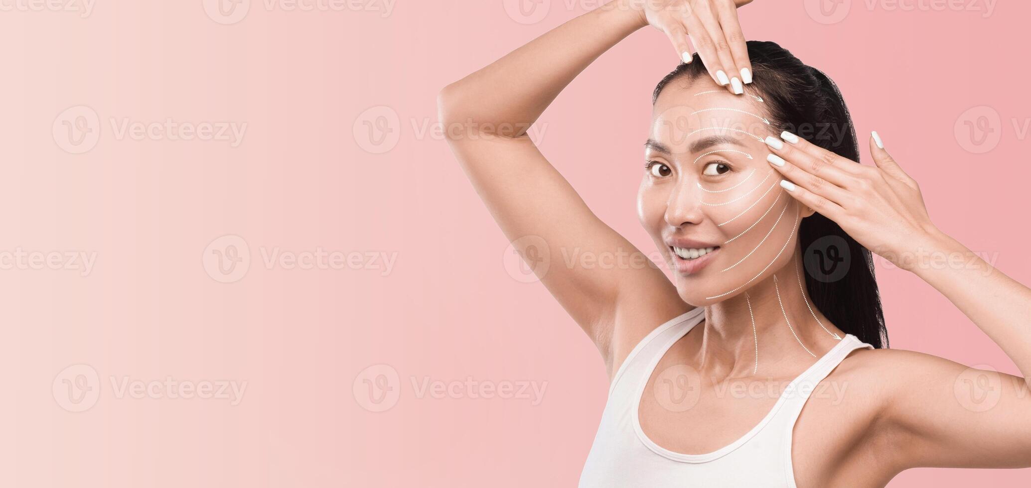 Asian woman is applying a facial mask as part of her skincare routine, surrounded by a calm, pink backdrop. The setting promotes relaxation and beauty, highlighting the pampering experience. photo
