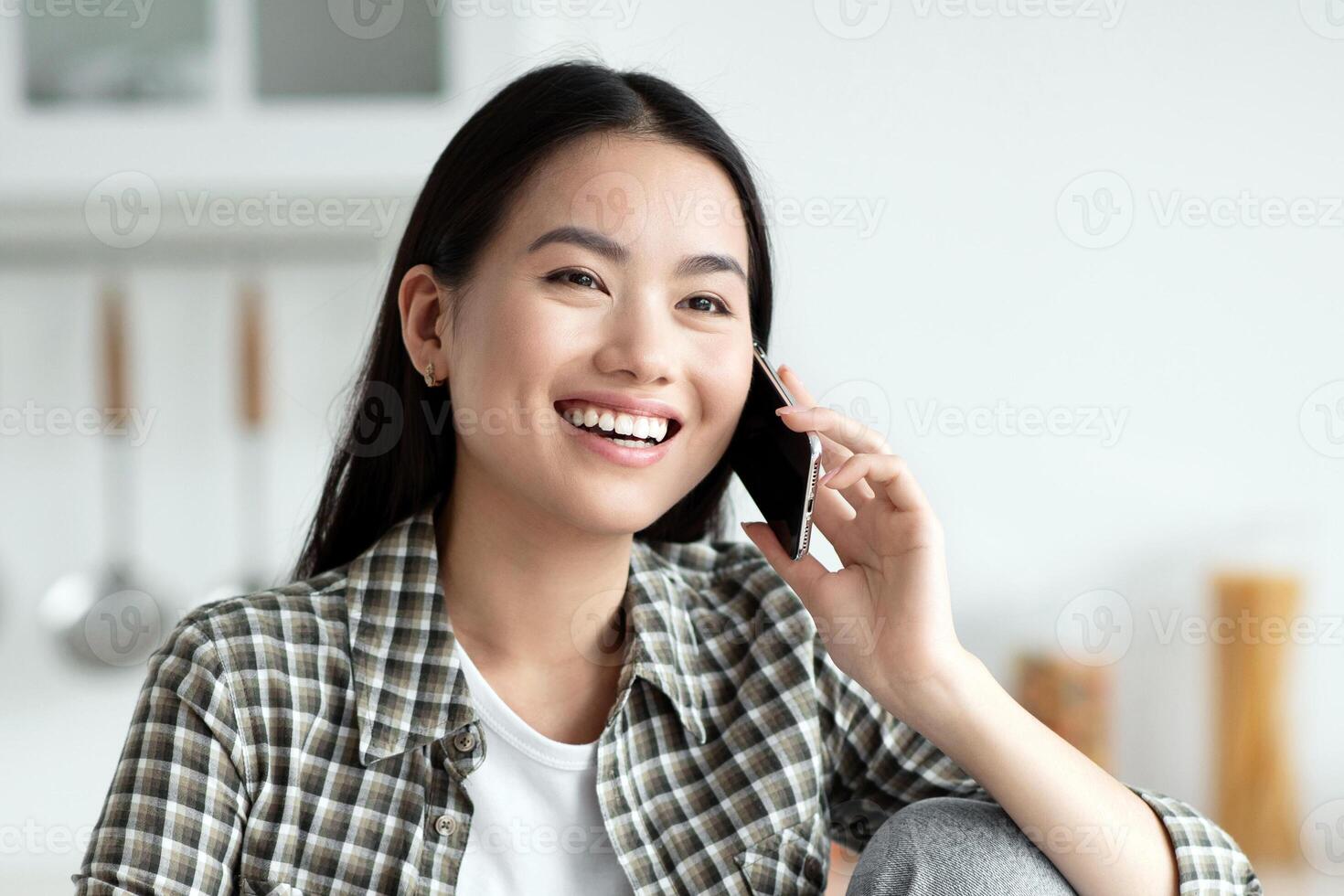 Young asian lady having healthy breakfast and talking on phone, smiling and looking at copy space. Cheerful woman sitting at kitchen, eating oatmeal and chatting with girlfriend or lover photo