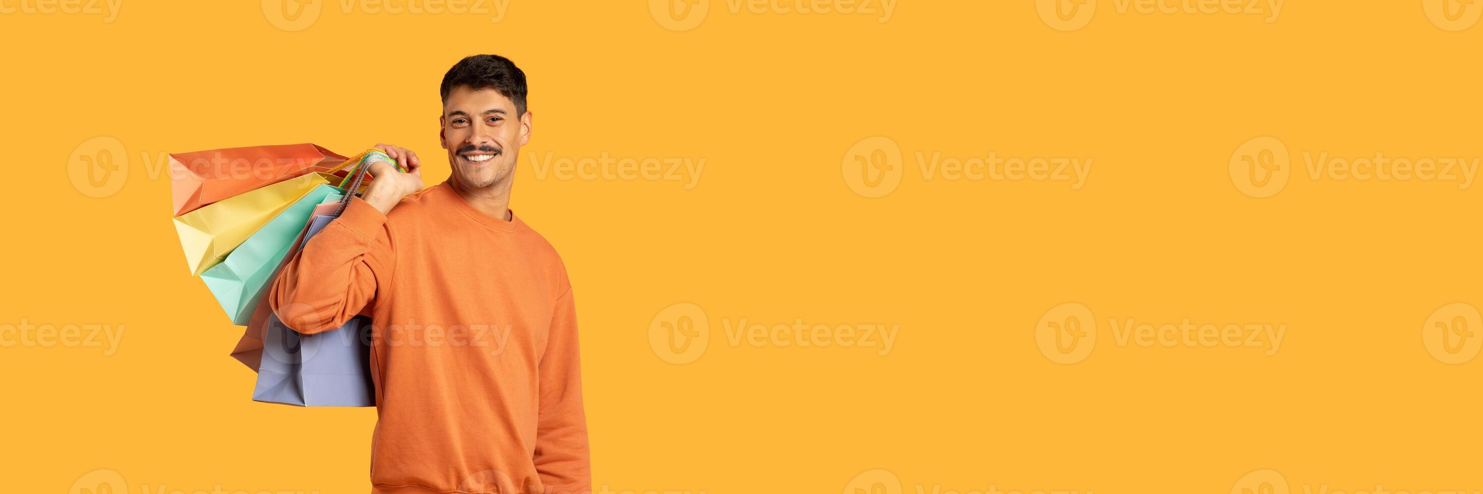 A millennial guy with a moustache carrying colorful shopping bags against an isolated yellow background, looking funny and content photo