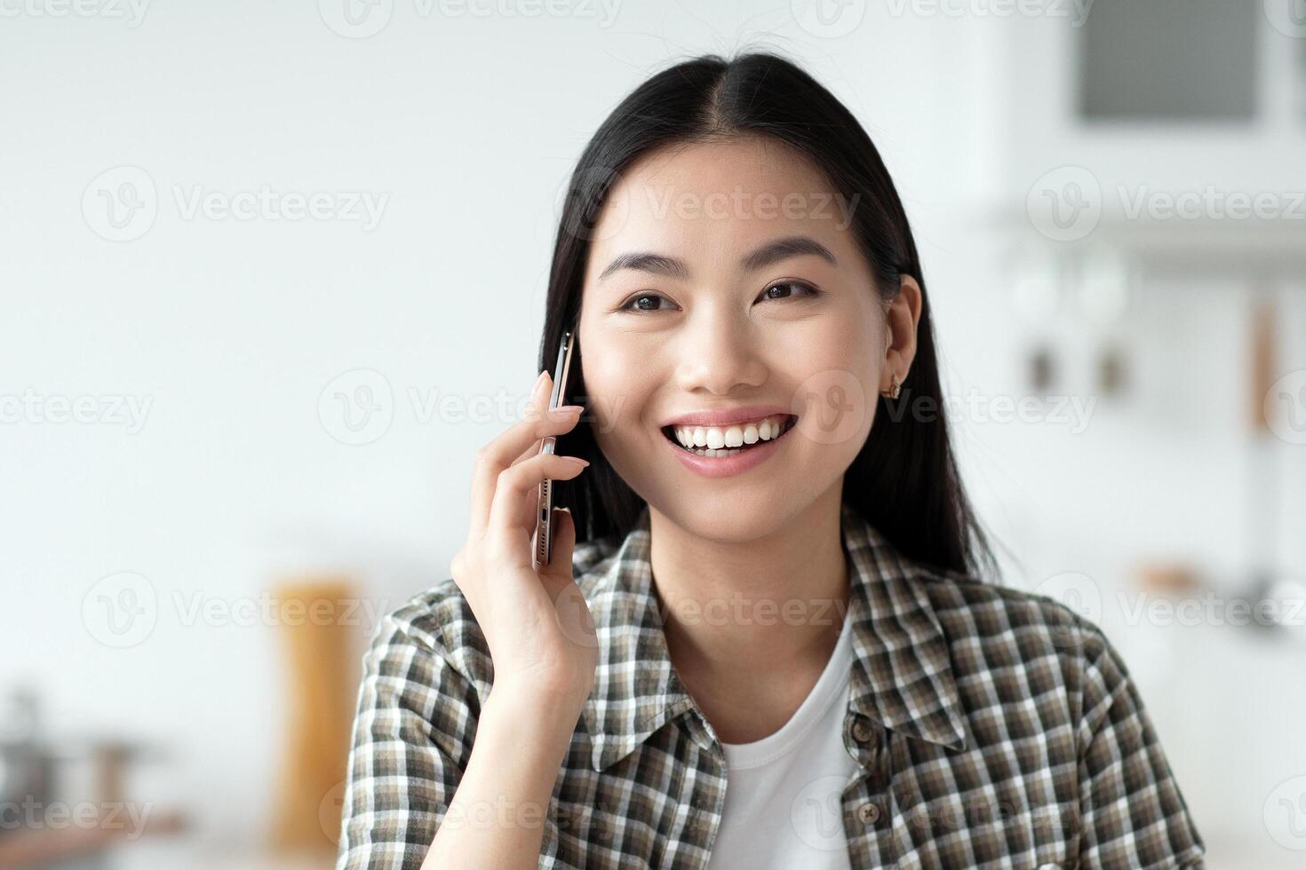 Cheerful young asian lady having healthy breakfast and talking on phone, smiling and looking at copy space. Pretty woman sitting at kitchen, eating oatmeal and calling girlfriend or lover photo