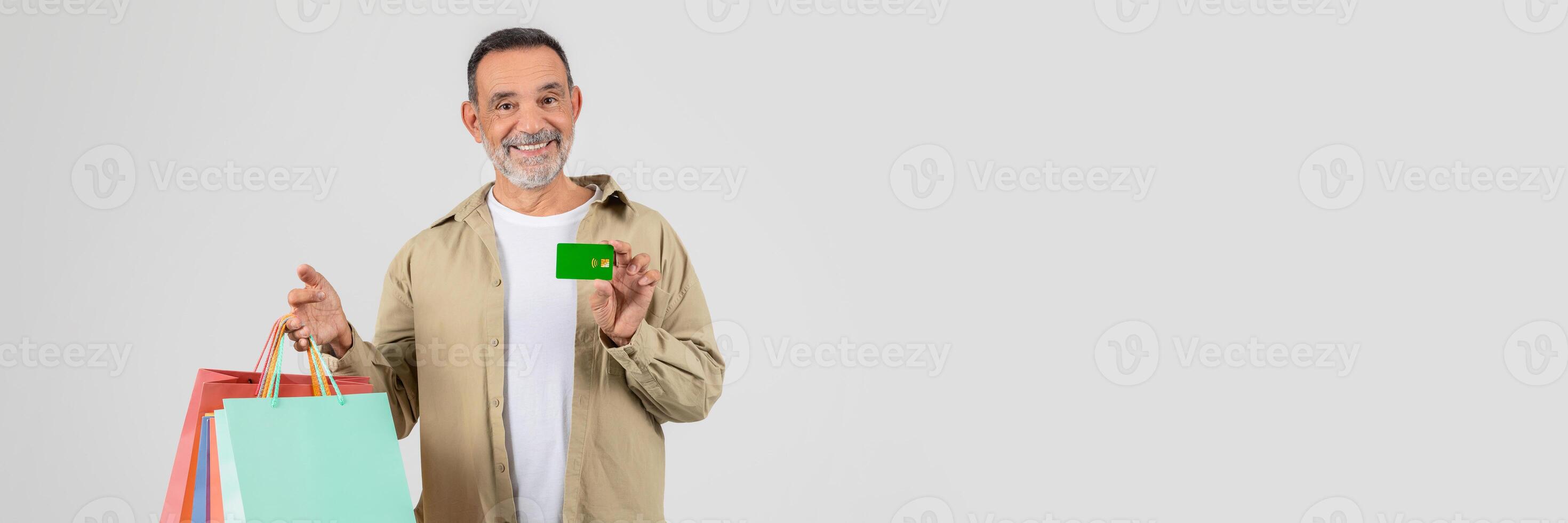 A senior man standing holding shopping bags in one hand and a credit card in the other. He appears to be in a shopping area, possibly a mall, ready to make purchases. photo