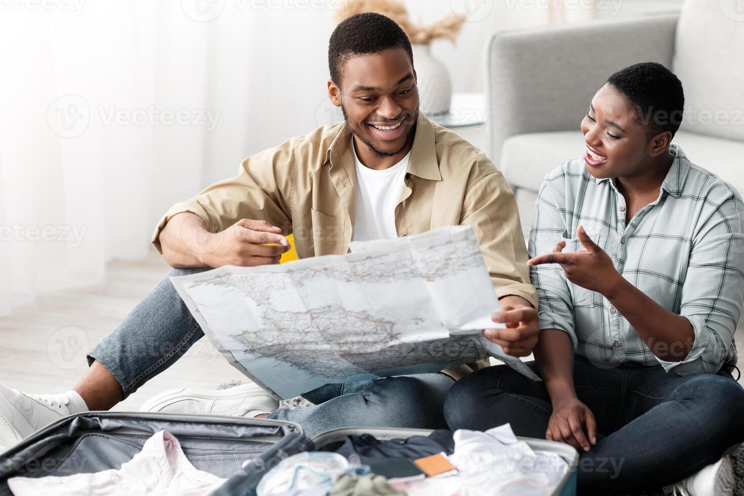 Travel Destination Choice And Navigation. Black Spouses Holding Touristic Map Choosing Country For Next Vacation Trip Together Sitting With Suitcase On Floor At Home. Tourism And Traveling Concept photo