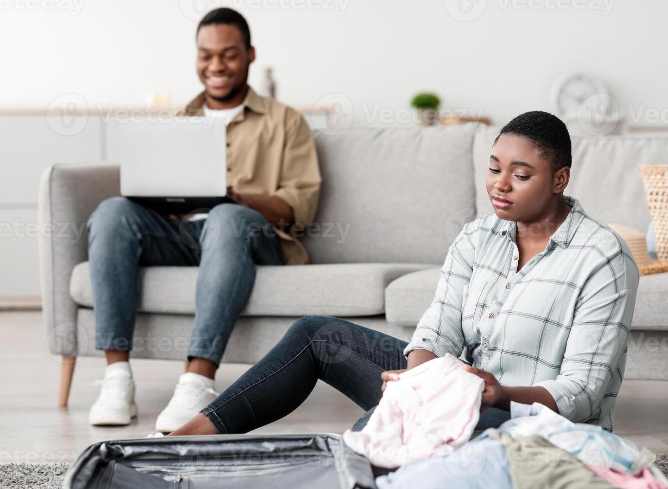Frustrated Black Wife Packing Travel Suitcase While Carefree Husband Using Laptop Computer Browsing Internet At Home. Selective Focus On Unhappy African American Lady Dreaming About Vacation photo
