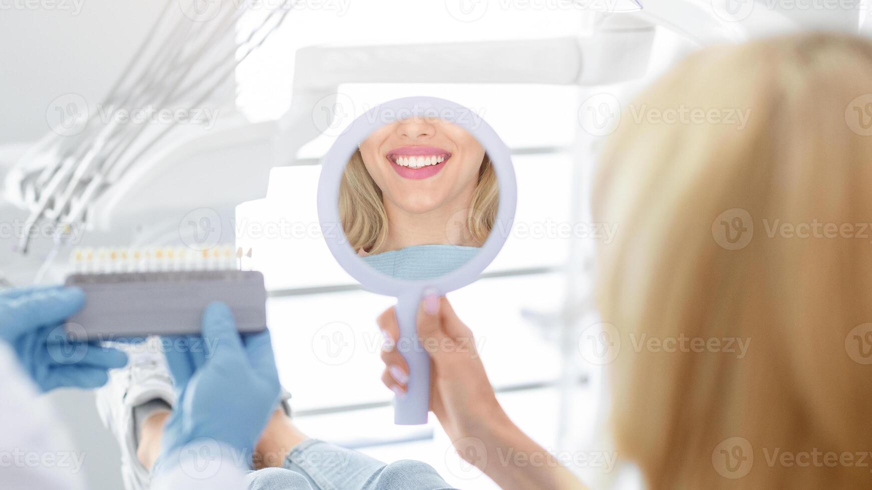 Dentist in blue medical gloves applying sample from tooth enamel scale to unrecognizable woman patient white teeth reflection in mirror to pick up right shade, teeth bleaching procedure, cropped photo
