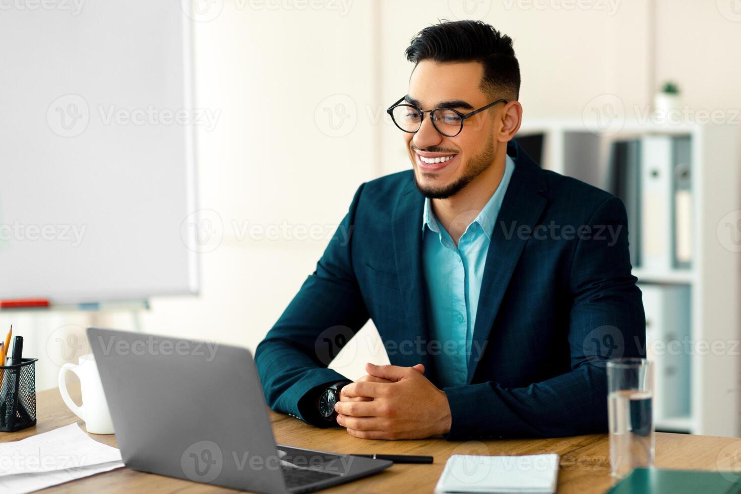 Confident young Arab entrepreneur in formal suit using laptop computer at his desk, having virtual meeting at office. Millennial businessman using pc for online job, communicating remotely photo