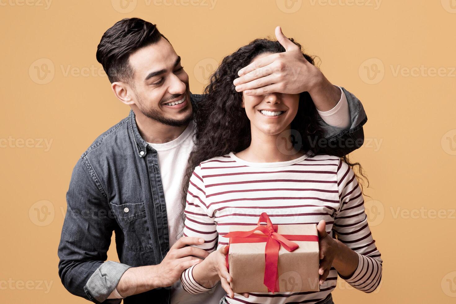 Surprise Gift. Romantic arab man covering his girlfriend's eyes and giving present, caring man greeting wife with birthday, wedding anniversary or valentines day while standing on yellow background photo