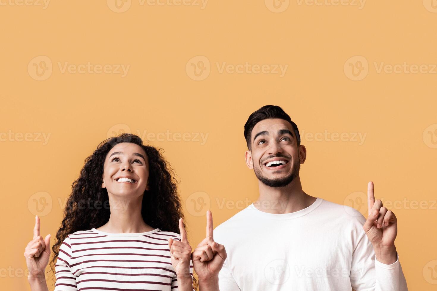 Check This. Joyful Young Arab Couple Pointing Up At Copy Space Above Their Heads, Cheerful Middle Eastern Man And Woman Showing Free Place On Yellow Background For Your Offer Or Advertisement photo