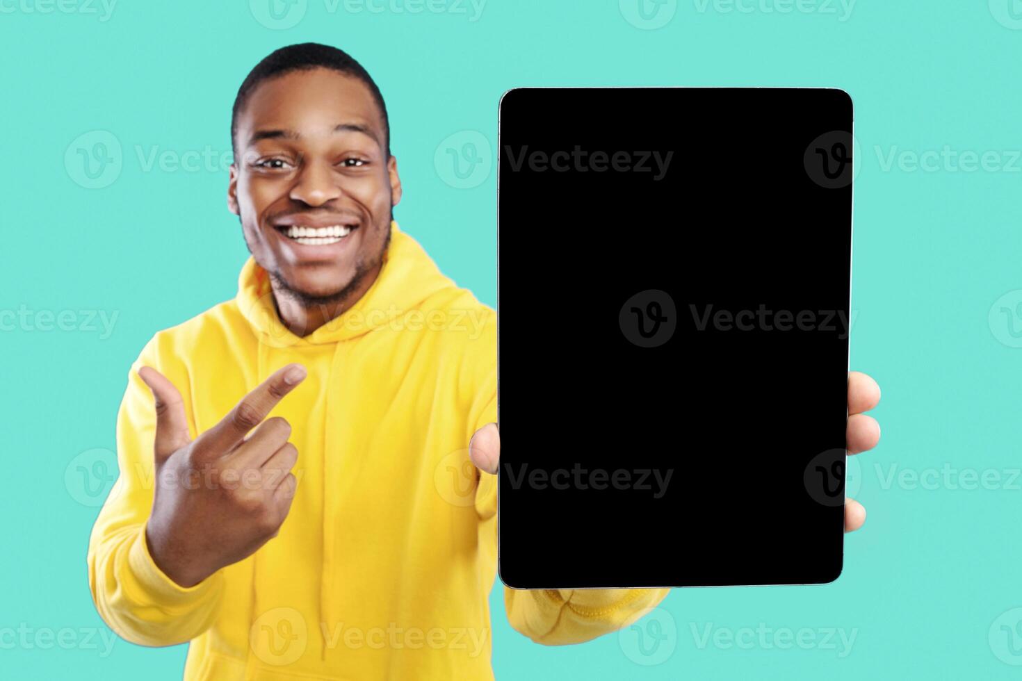 Black Guy Showing Digital Tablet With Blank Screen Recommending New App Or Great Website, Posing Pointing Finger Over Blue Background In Studio. Selective Focus, Mockup photo