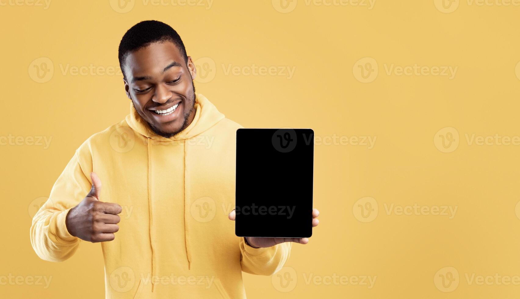 African American Guy Showing Tablet Computer Blank Screen Gesturing Thumbs Up Approving Website Posing Over Yellow Background. Studio Shot. People And Gadgets Concept. Mockup photo