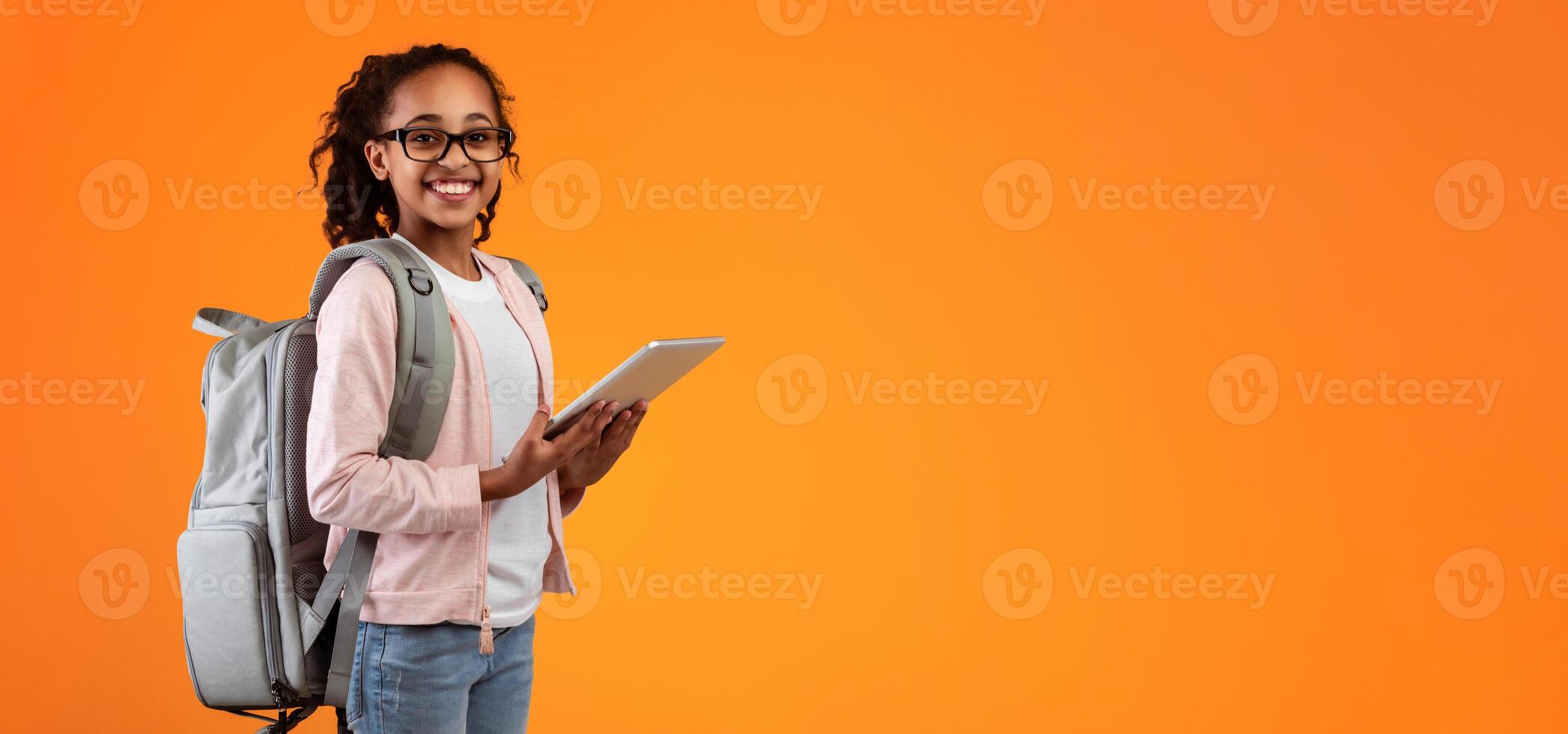 Online Classes Concept. Smiling teenage pupil wearing glasses and backpack holding and using digital tablet for studying standing on yellow orange studio wall posing at camera, free copy space photo