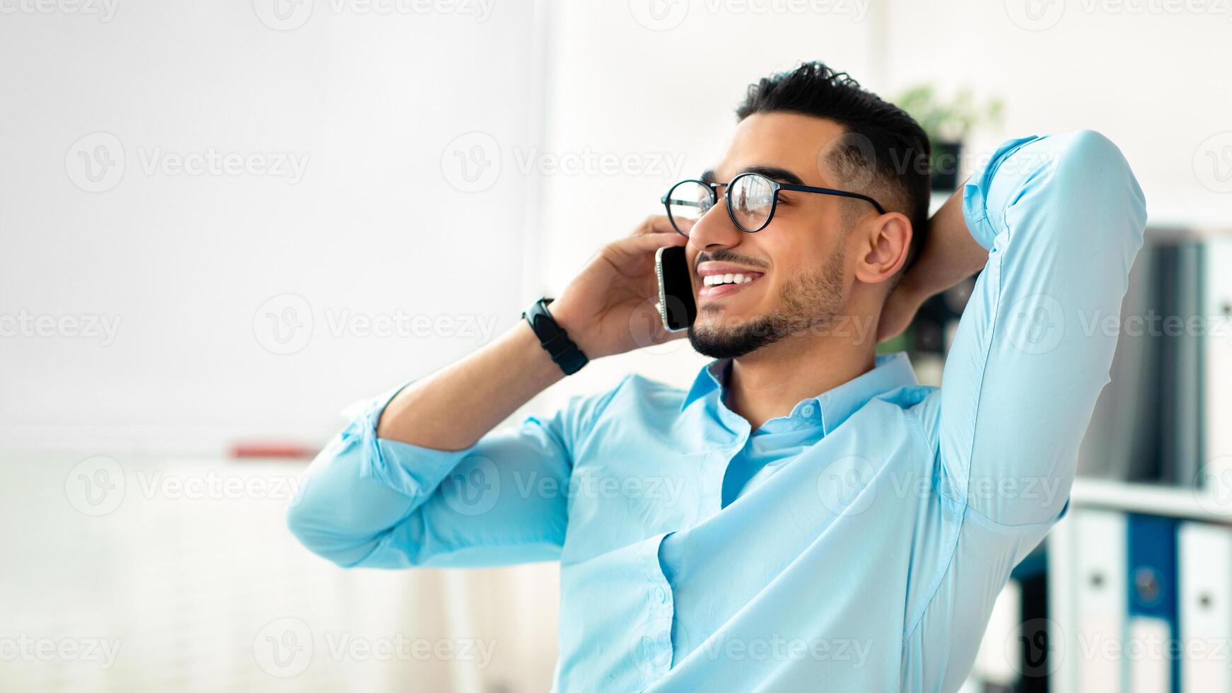 Handsome young Arab man in office wear talking on mobile phone at workplace. Attractive guy entrepreneur having phone conversation with client or business partner, indoors photo