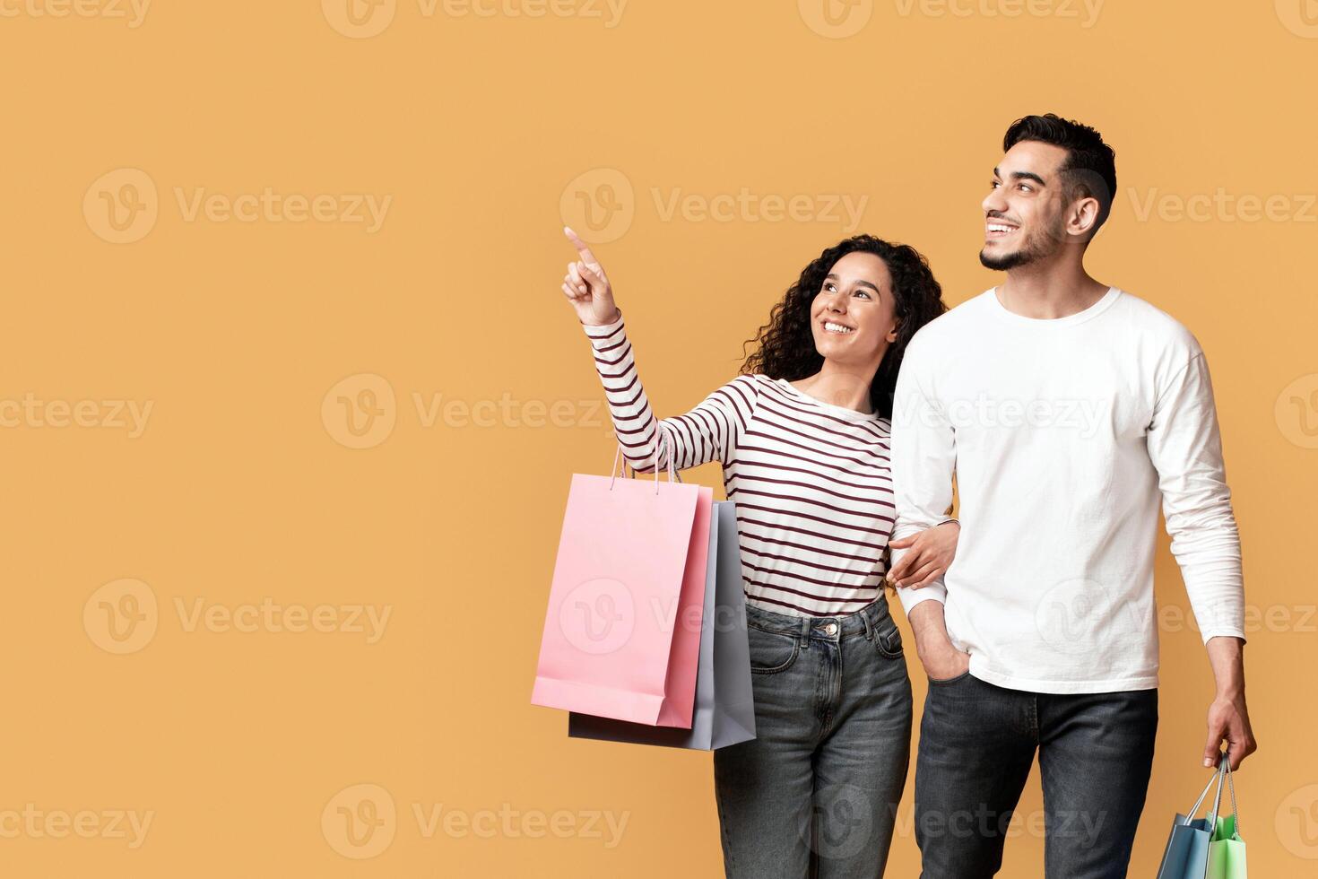 Seasonal Discounts. Smiling Arab Spouses Walking With Shopping Bags And Pointing Aside On Yellow Background In Studio, Happy Middle Eastern Couple Enjoying Shopper Deals, Panorama With Copy Space photo