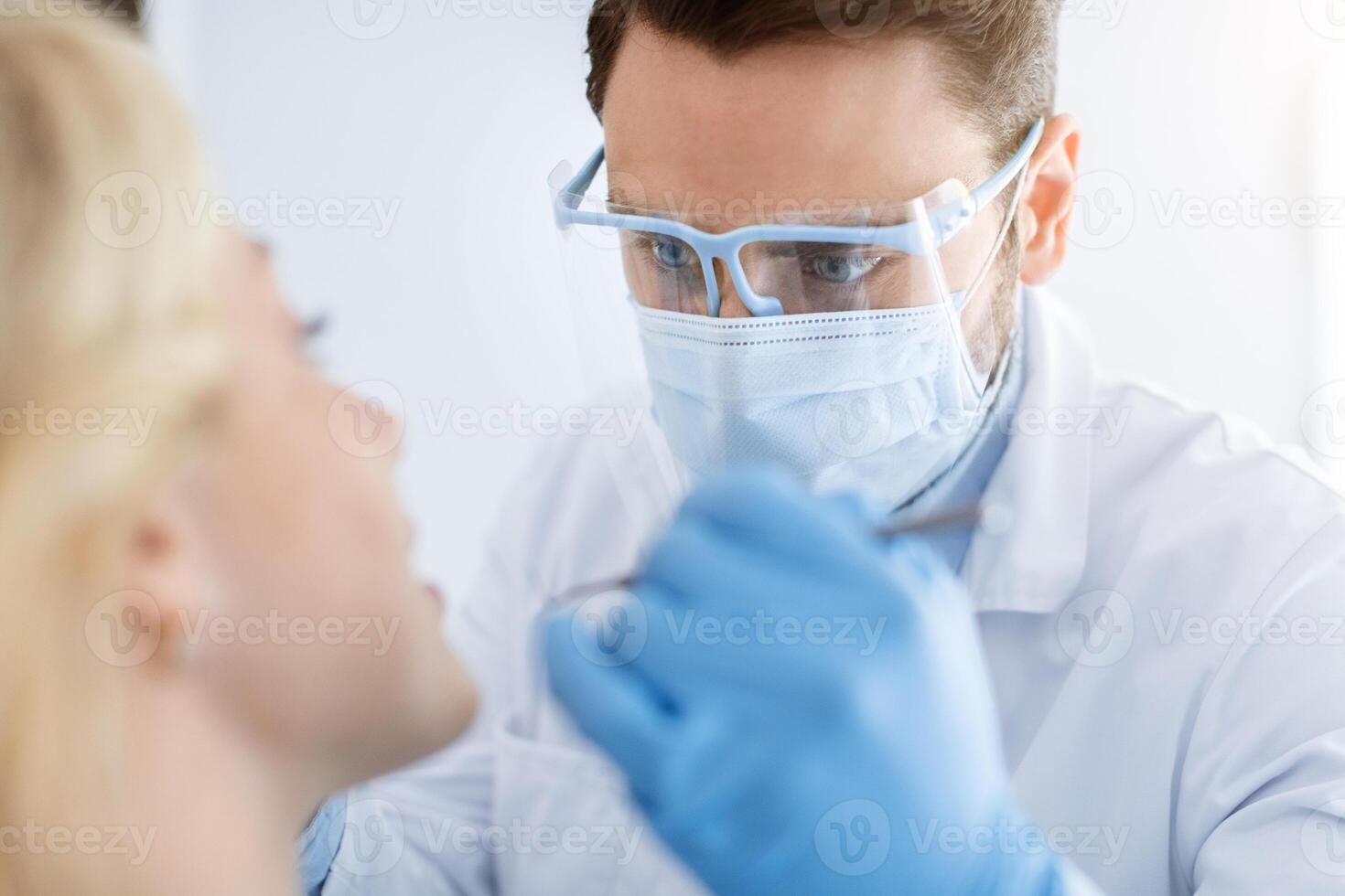 Closeup of young man dentist wearing medical face mask and protective glasses doing tratment or checkup for young blonde woman, wearing blue rubber gloves and holding dental tools photo