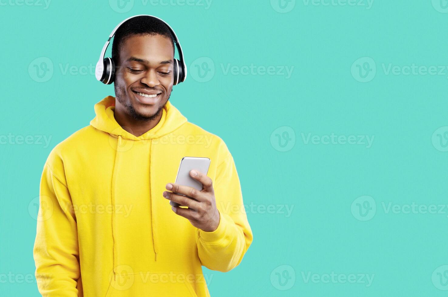 African American Guy In Headphones Using Smartphone Texting And Listening To Music Standing Over Blue Studio Background, Wearing Yellow Hoodie. Great Musical App Concept photo