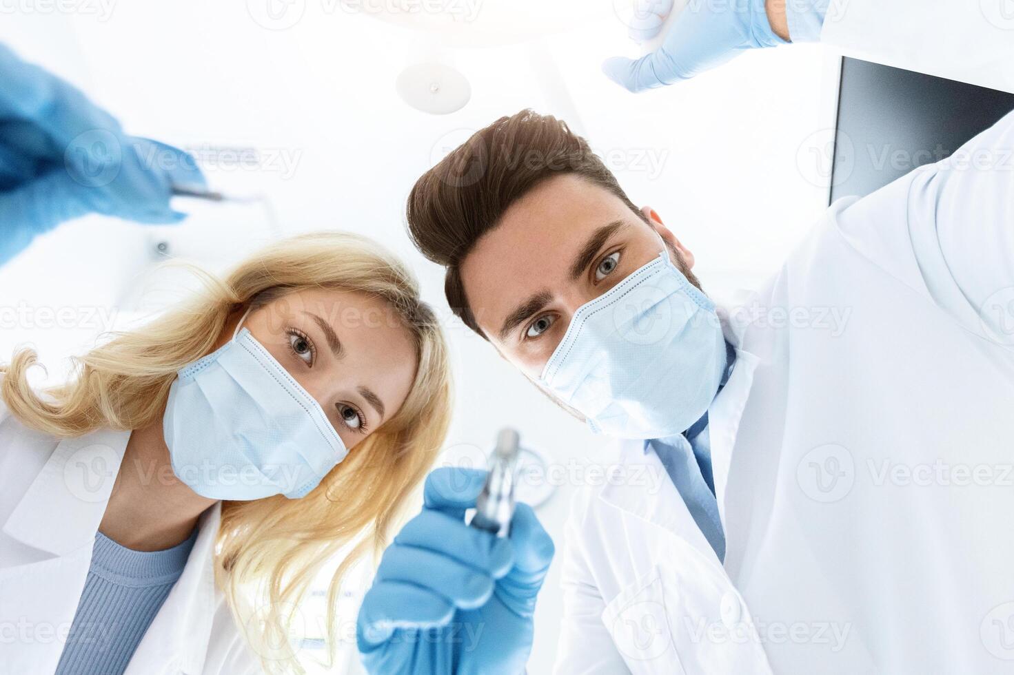 Two dentists young caucasian man and woman in face masks and rubber gloves doing treatment, turning on lamp, shot from dental chair. Modern stomatology, high class dental clinic concept photo