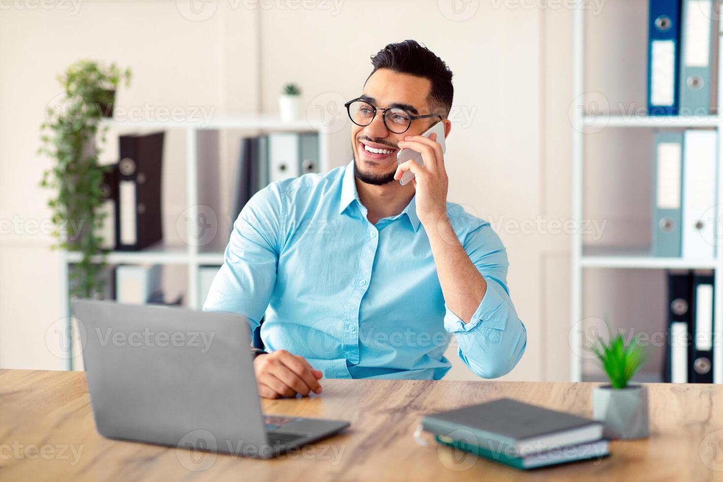 Handsome young Arab man in formal wear talking on smartphone, enjoying conversation at workplace. Attractive millennial entrepreneur speaking with customer while working on laptop at office photo