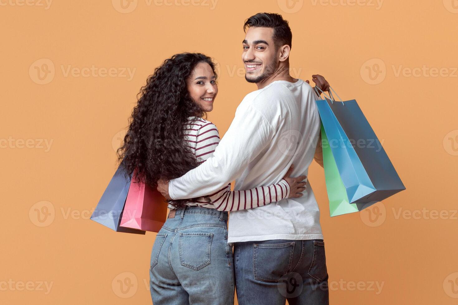 Family Shopping. Happy Arab Spouses Embracing And Holding Colorful Shopper Bags, Smiling Middle Eastern Couple Walking On Yellow Studio Background And Turning At Camera, Full Length, Copy Space photo