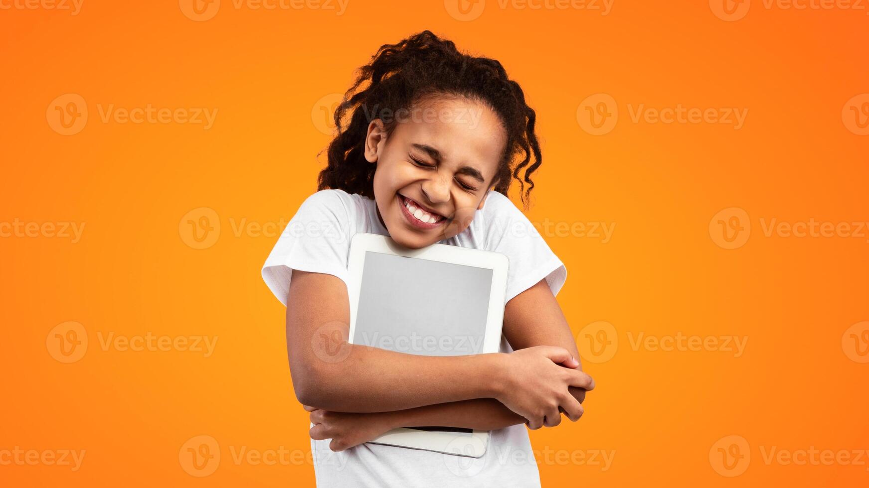 Technology Lover Concept. Happy black teenage girl hugging digital tablet with blank empty screen, holding pc tight near chest. Female geek posing isolated over yellow studio background, copy space photo