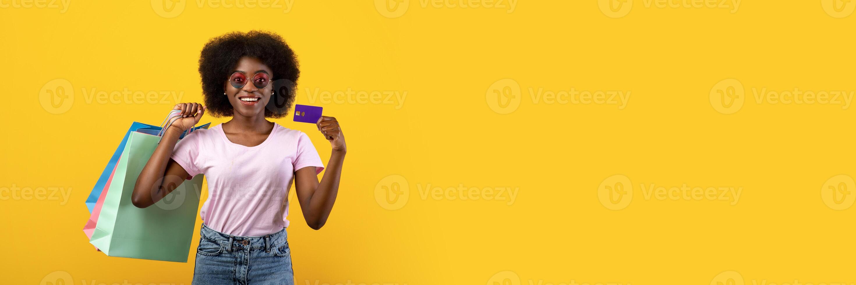 Easy shopping. Excited african american woman holding credit card and shopper bags, posing over yellow studio background. Happy lady carrying purchases, got cashback bonus photo