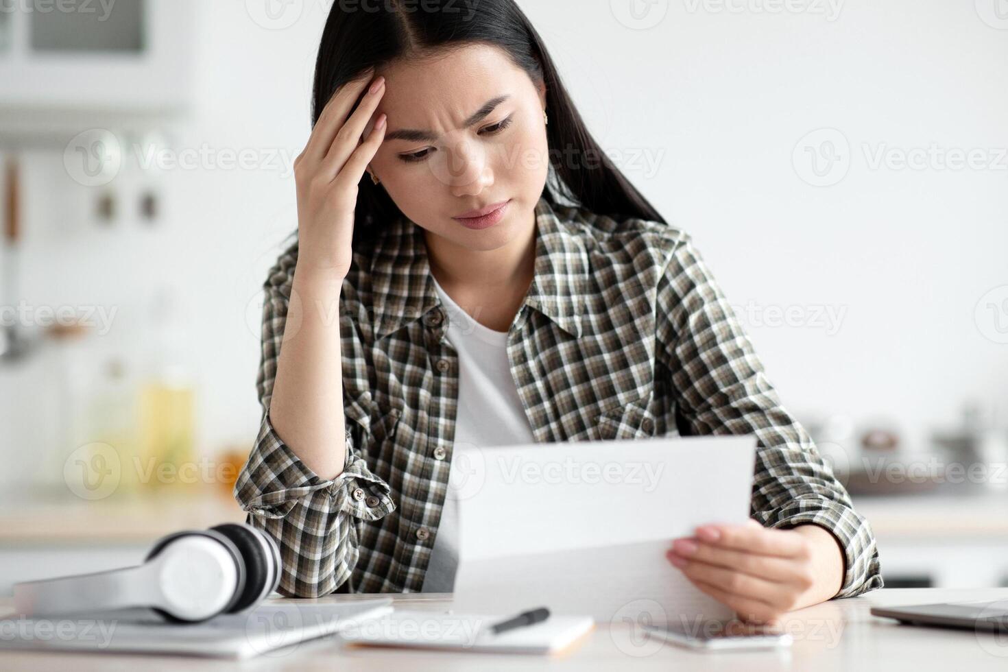 Upset young asian woman sitting at table with modern laptop in kitchen, reading letter and touching her forehead, copy space. Sad lady holding document or bills, got bad news, home interior photo