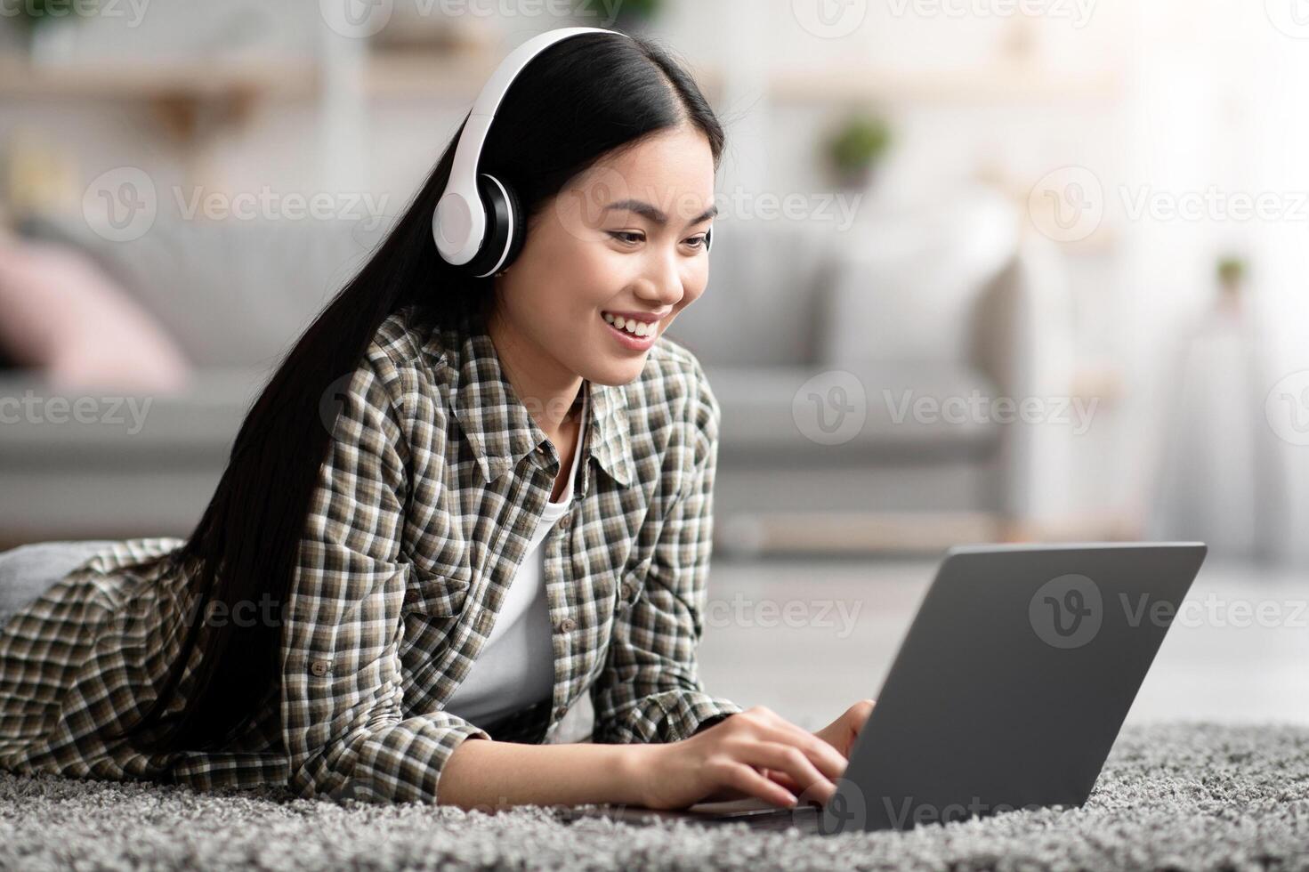 Joyful asian lady using wireless headset and laptop while relaxing at home alone, laying on carpet in living room, playing games or watching movie online, copy space. Domestic entertainment photo