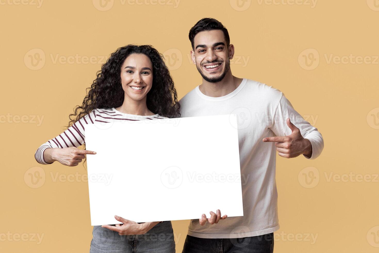 Place For Ad. Happy Arab Couple Holding And Pointing At Blank White Placard In Their Hands, Smiling Young Middle Eastern Man And Woman Demonstrating Copy Space For Advertisement, Mockup photo