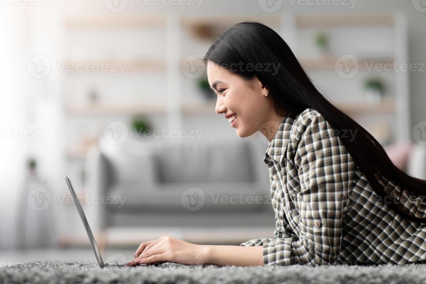Side view of asian young woman using laptop at home, laying on carpet in living room, typing on computer keyboard, chatting with lover or friends while resting at weekend, copy space photo