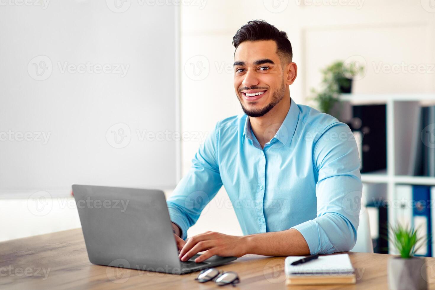 Attractive young Arab businessman using laptop pc at his desk in home office. Cheerful Eastern guy entrepreneur typing on computer keyboard, having in online conference at workplace photo