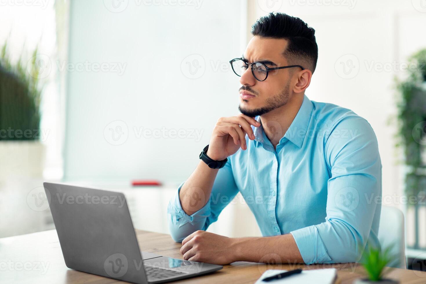 Troubled Arab businessman sitting at his desk, looking at laptop screen, solving work problem in modern office. Millennial guy entrepreneur suffering from work stress, facing project deadline photo