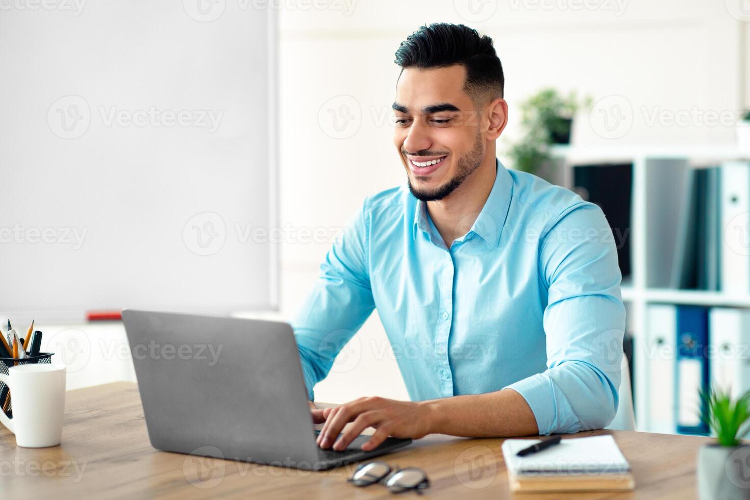 Handsome young Arab businessman working with laptop computer at his desk in office. Happy guy entrepreneur typing on pc keyboard, checking email, taking part in online meeting at workplace photo