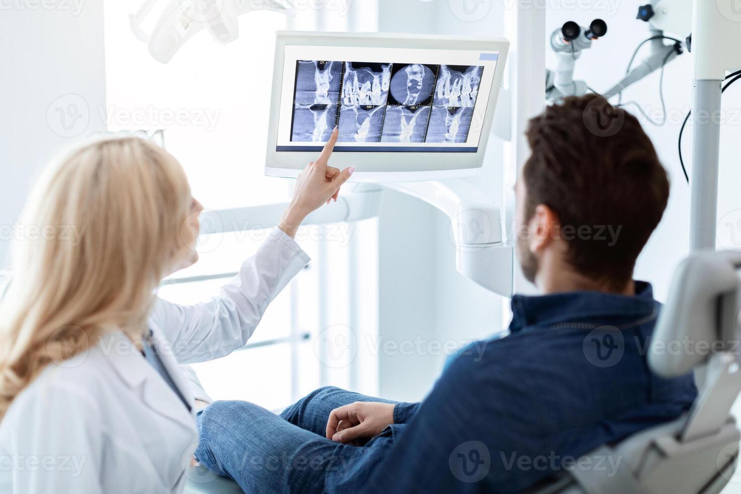 Modern dental clinic concept. Blonde woman dentist having conversation with patient in dental chair, poitning at LED display, showing results of treatment or explaining procedure photo