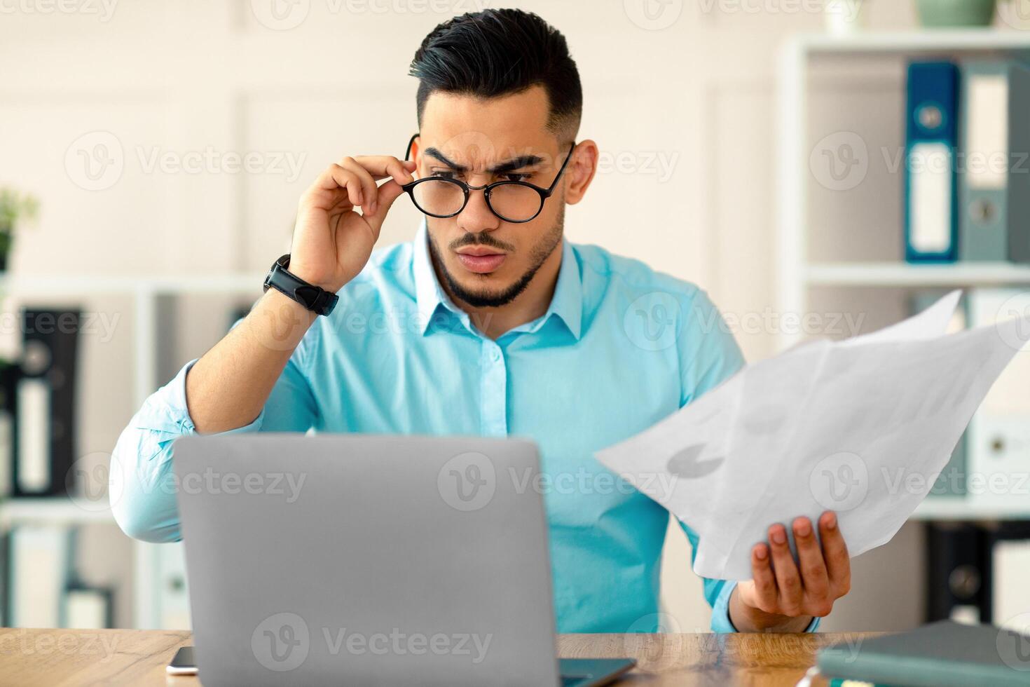 Stressed young Arab employee using laptop, holding documents, having problem with work task, making mistake in business project at office. Millennial guy having trouble meeting deadline photo