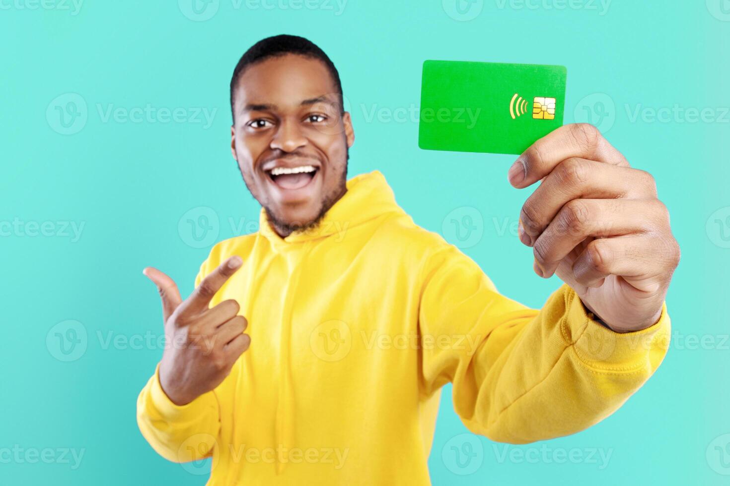Excited African American Guy Showing Green Credit Card Pointing Finger At It Smiling To Camera Posing Over Blue Studio Background. Guy Advertising Great Bank Service. Money And Finance photo