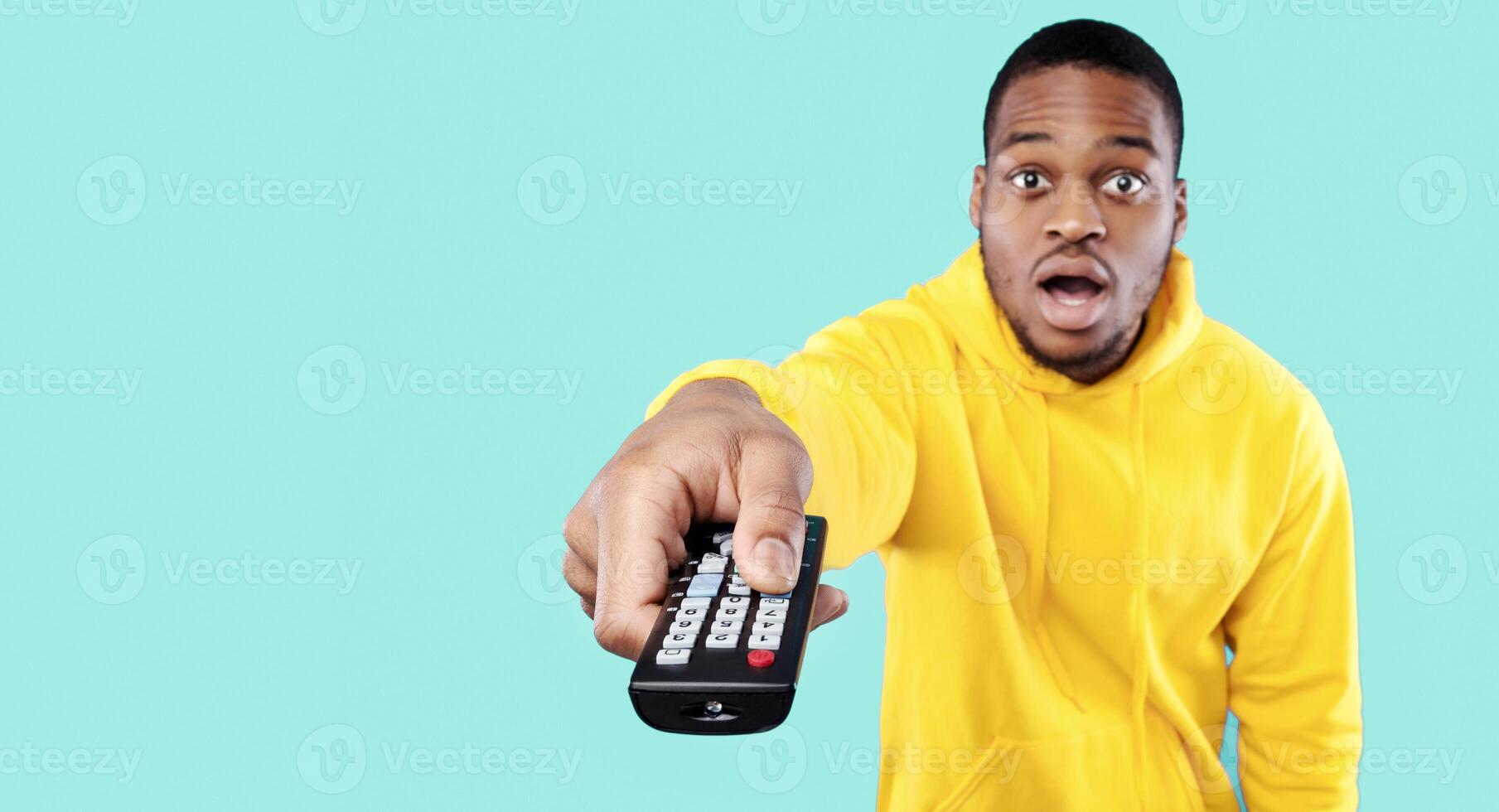 Shocked African Man Watching Television Pointing TV Remote Controller To Camera Switching Channels With Opened Mouth Over Blue Background. Studio Shot. Shocking TV Content. Selective Focus photo
