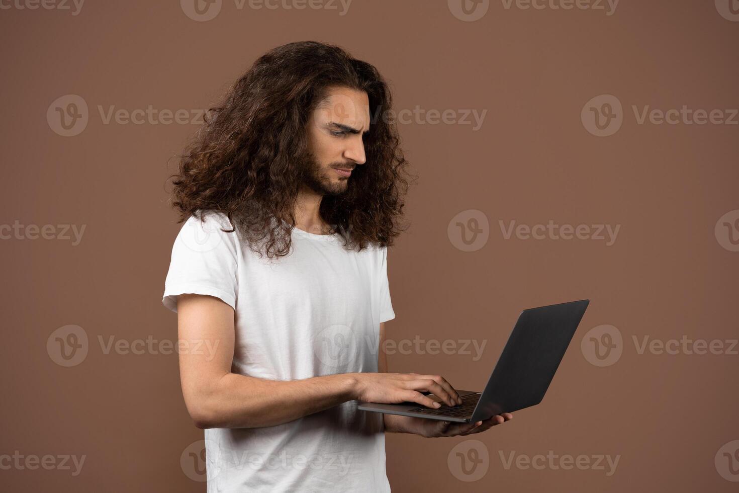 A young man with long curly hair stands alone, intently using a laptop. He is dressed casually in a white t-shirt, set against a warm brown background that enhances his concentration and lifestyle. photo